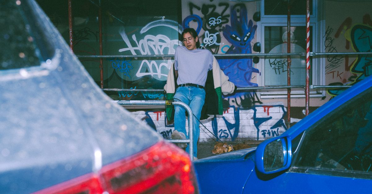 Jovem adulto posando perto de um muro grafitado em Berlim, cercado por carros estacionados.