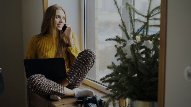 Young woman sitting by the window using a laptop and talking on a phone, enjoying a cozy indoor setting.