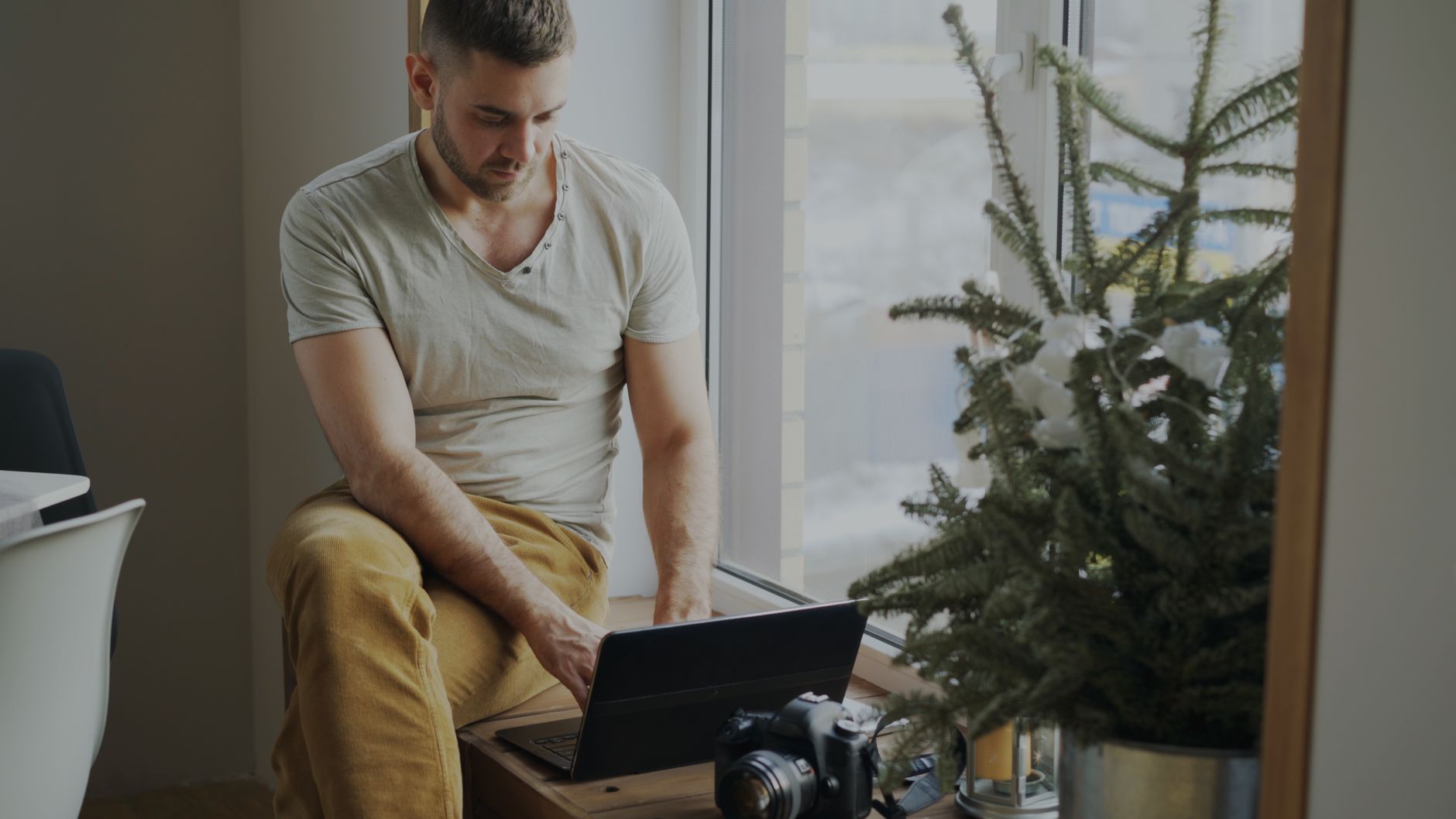 Casual man using laptop indoors by a window, with camera and plant nearby.