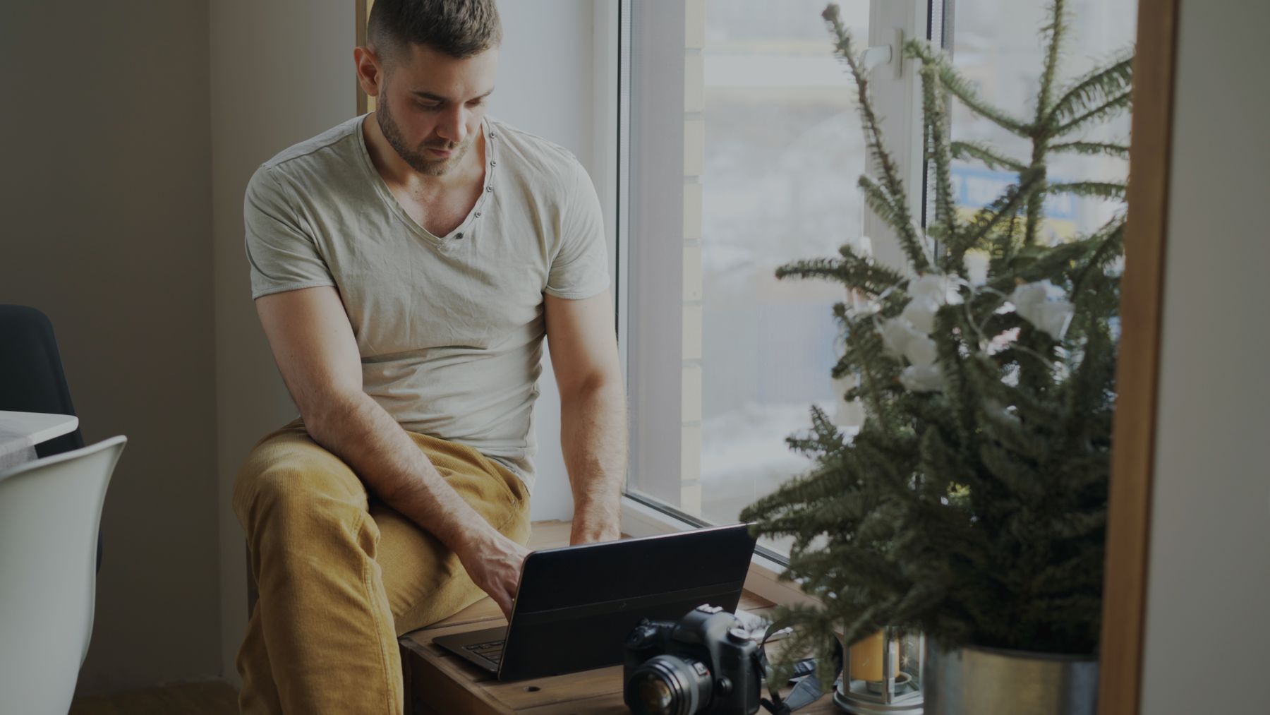 A man working on a laptop by a window with a camera and plant nearby, photo by Vitaly Gariev on Pexels