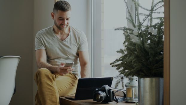 Young man sitting by the window using smartphone, laptop nearby with camera.