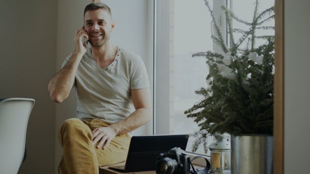 Smiling man sitting by window on phone, with laptop and camera.