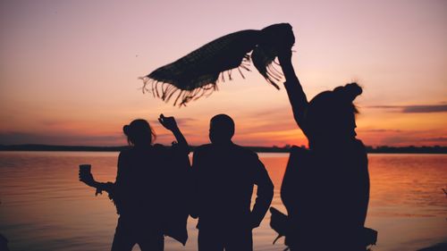Free Three friends silhouetted against a vibrant sunset by a tranquil lake, enjoying a moment of freedom. Stock Photo