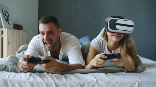 Free Young couple enjoying video games with controllers and VR headset on bed indoors. Stock Photo