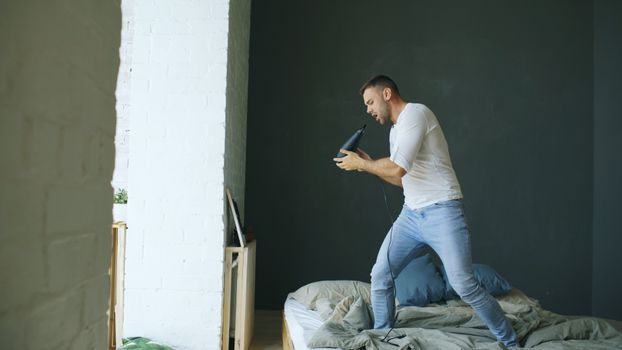Man humorously sings into a hairdryer while standing on a messy bed in a stylish bedroom.