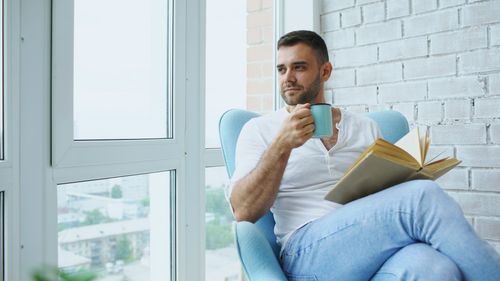 Free Man enjoys a quiet moment with coffee and a book in a cozy chair by the window. Stock Photo
