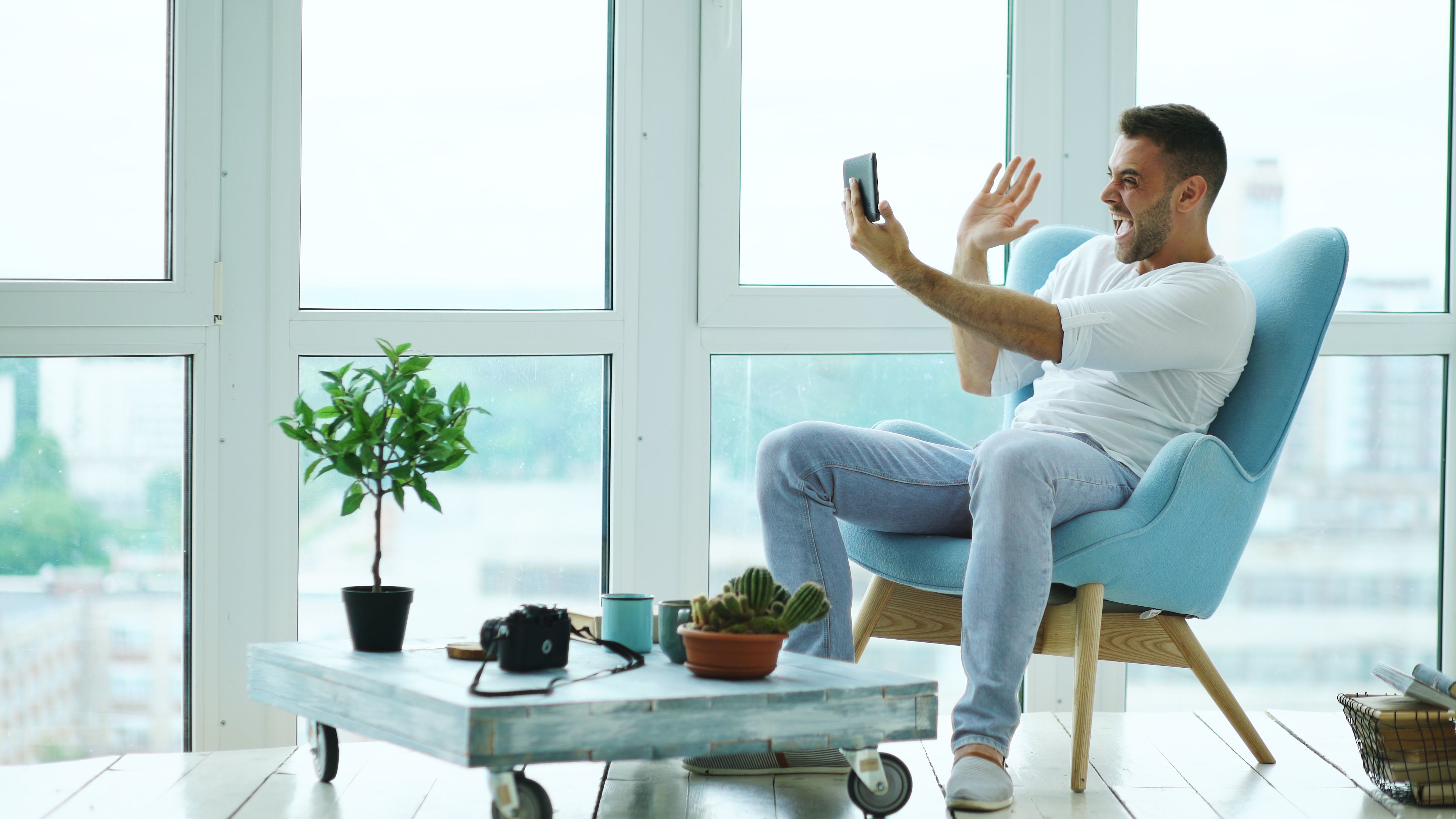 Joyful man waving during a video call on a tablet in a stylish, bright living room with large windows.