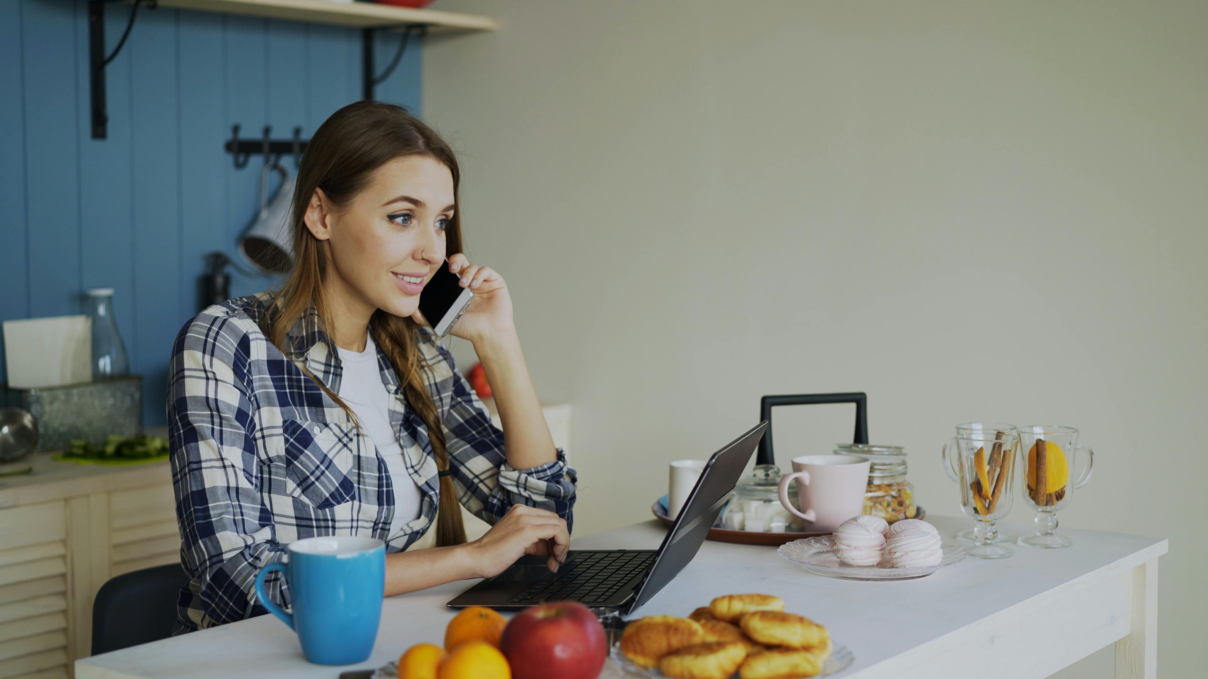 Young woman multitasking on phone and laptop in a cozy kitchen setting.