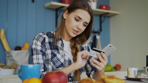 Woman in plaid shirt using credit card with smartphone in cozy kitchen setting.