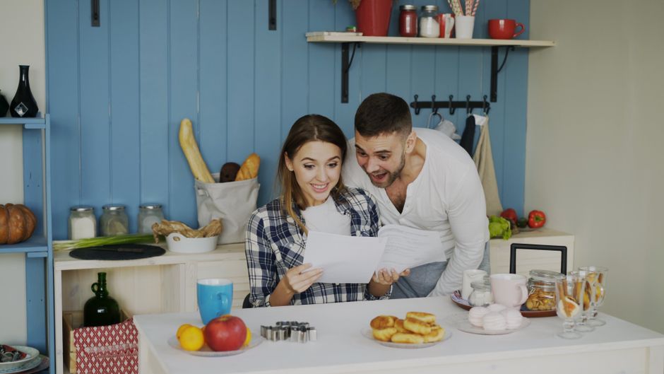 A cheerful couple in a kitchen excitedly reading documents, surrounded by food and kitchen items.