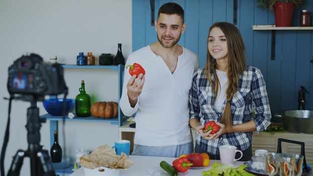 A couple prepares to film a cooking video while holding bell peppers in a stylish kitchen.