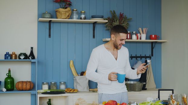 Smiling man holds coffee and checks phone in stylish kitchen setting.