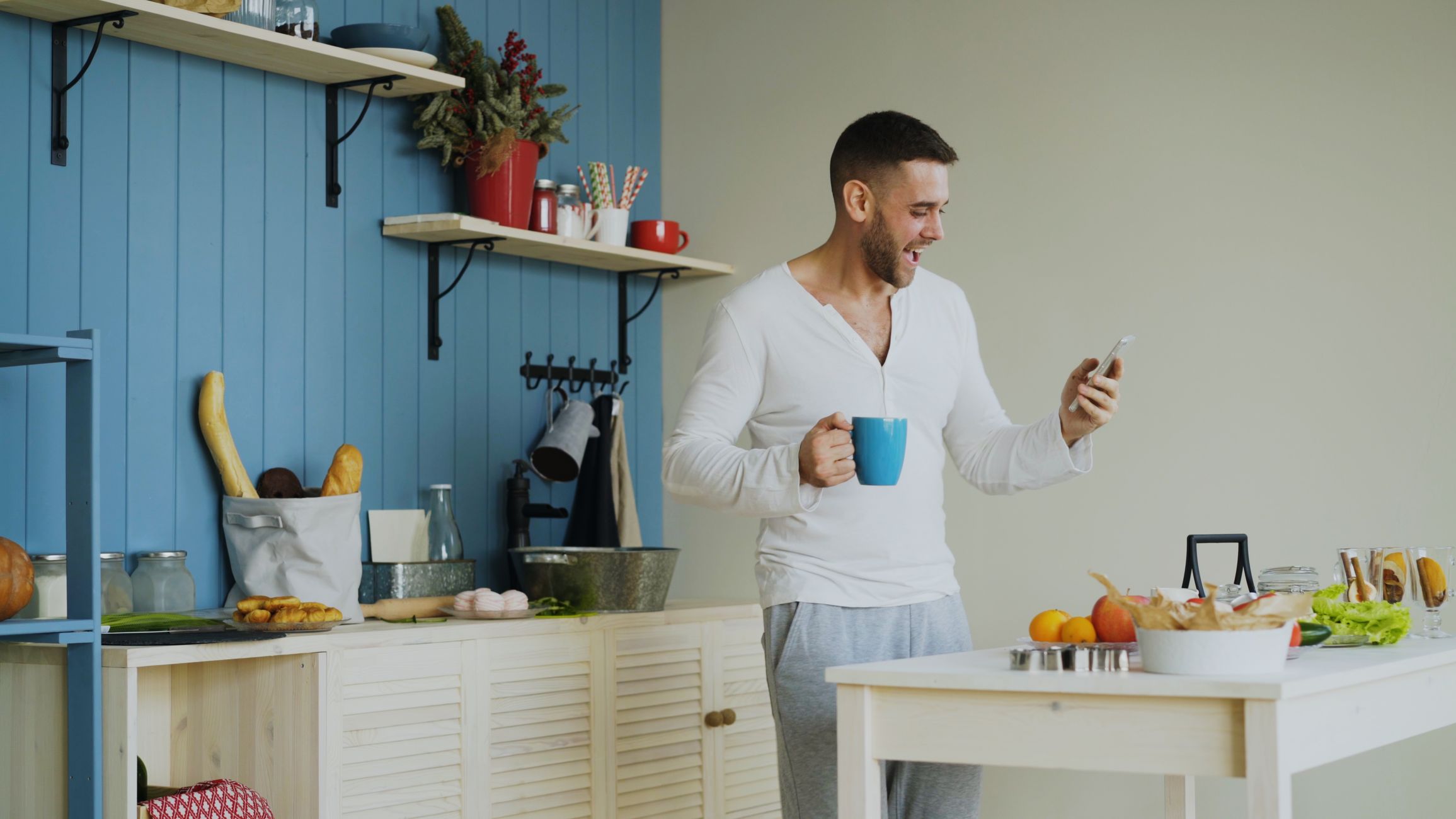 Smiling man holds a mug and phone in a bright modern kitchen setting.