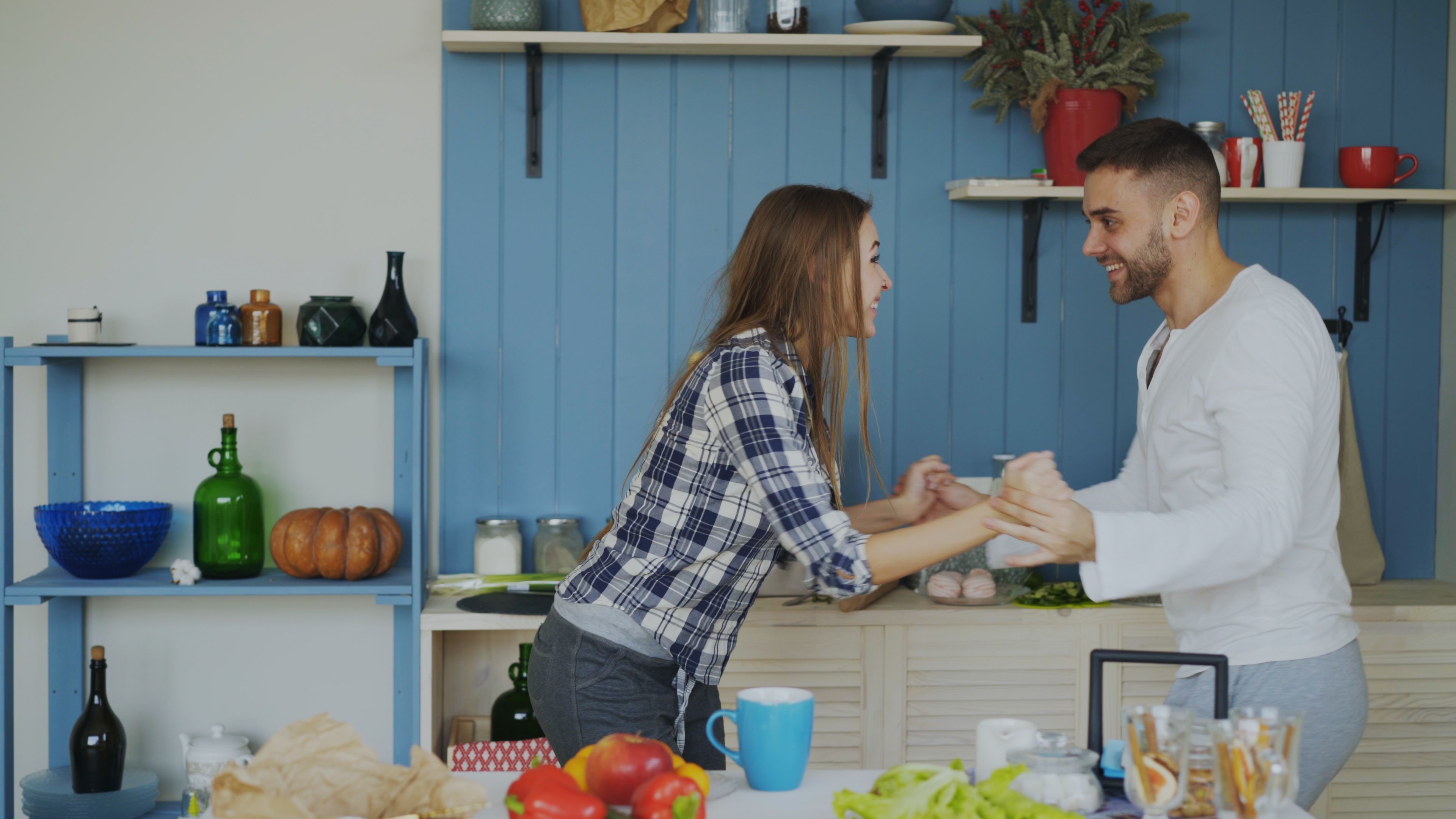 Young couple smiling and holding hands in a cozy kitchen setting, surrounded by food and decor.