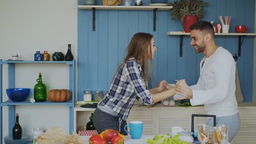 Free Young couple smiling and holding hands in a cozy kitchen setting, surrounded by food and decor. Stock Photo