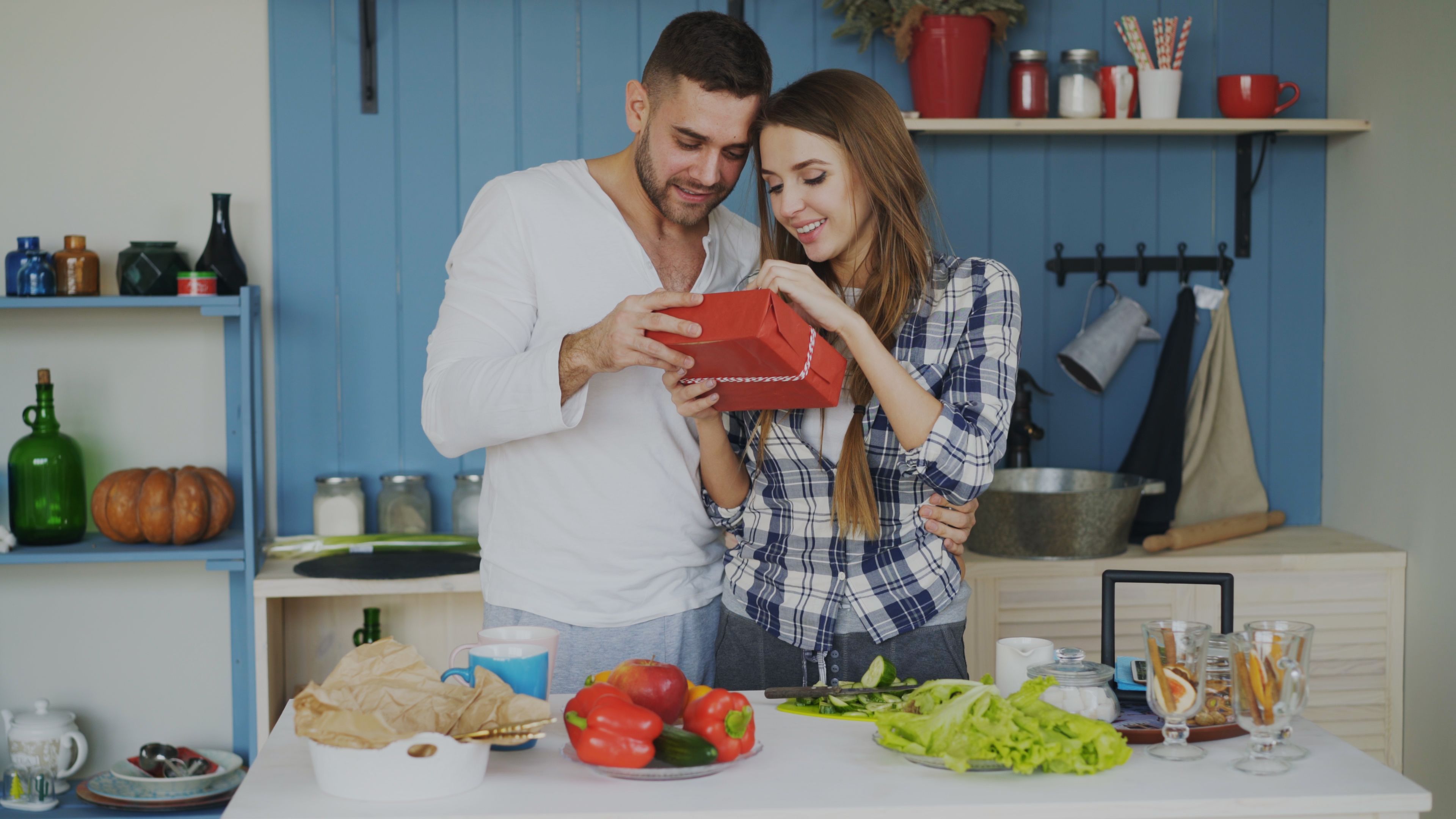 Casal feliz trocando presente em uma cozinha acolhedora decorada com utensílios e alimentos variados.
