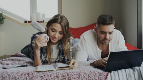 Free Couple lying on bed, reading book and using laptop, enjoying relaxed indoor time. Stock Photo