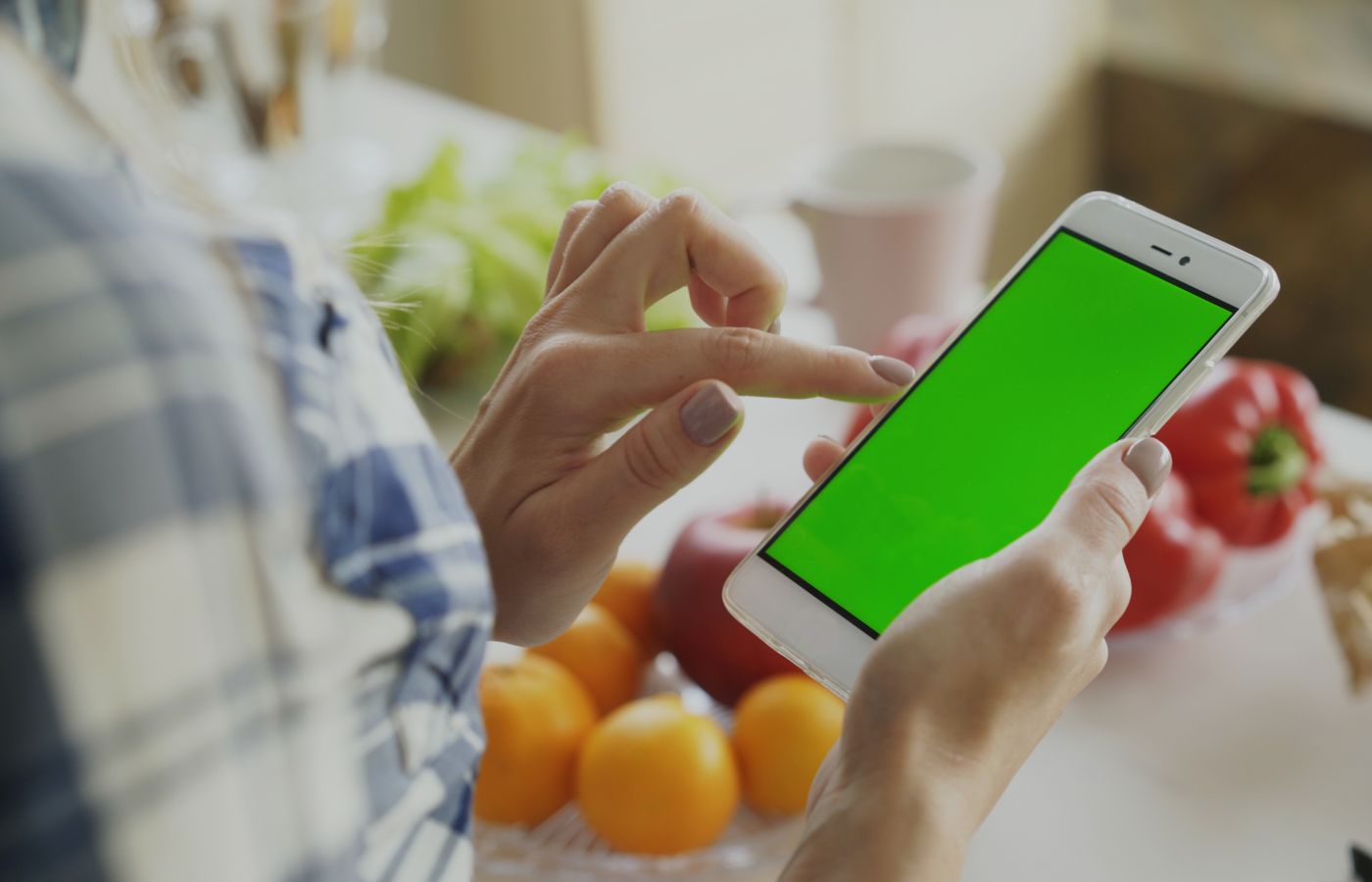 A person using a smartphone with a green screen surrounded by fresh fruits and vegetables in the kitchen.