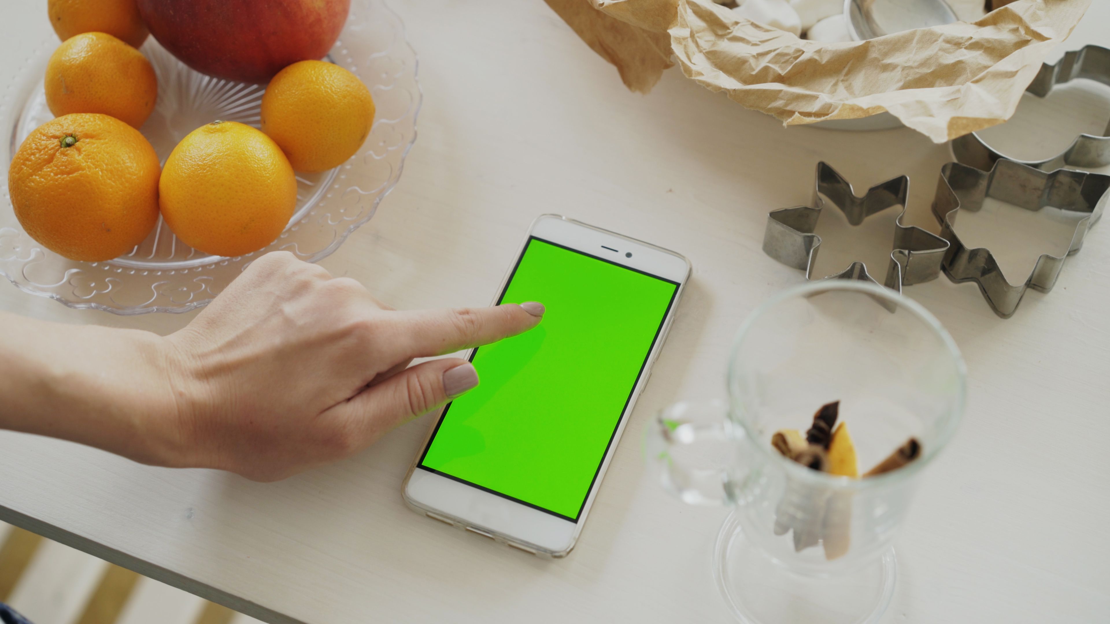 Hand touching a smartphone with green screen next to fresh fruit and cookie cutters on table.