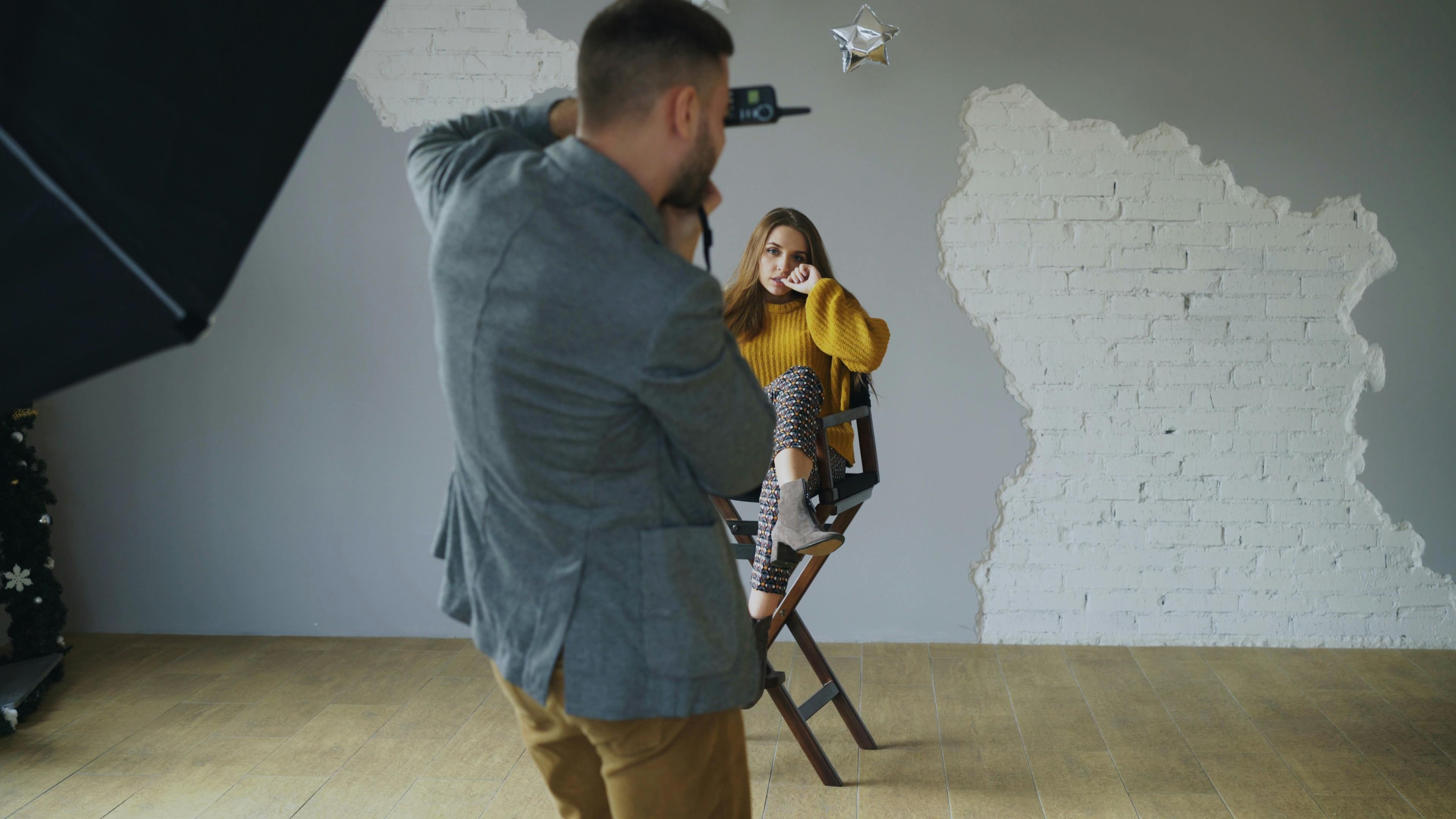 A photographer takes a photo of a stylish model seated on a chair in a studio with a textured wall.