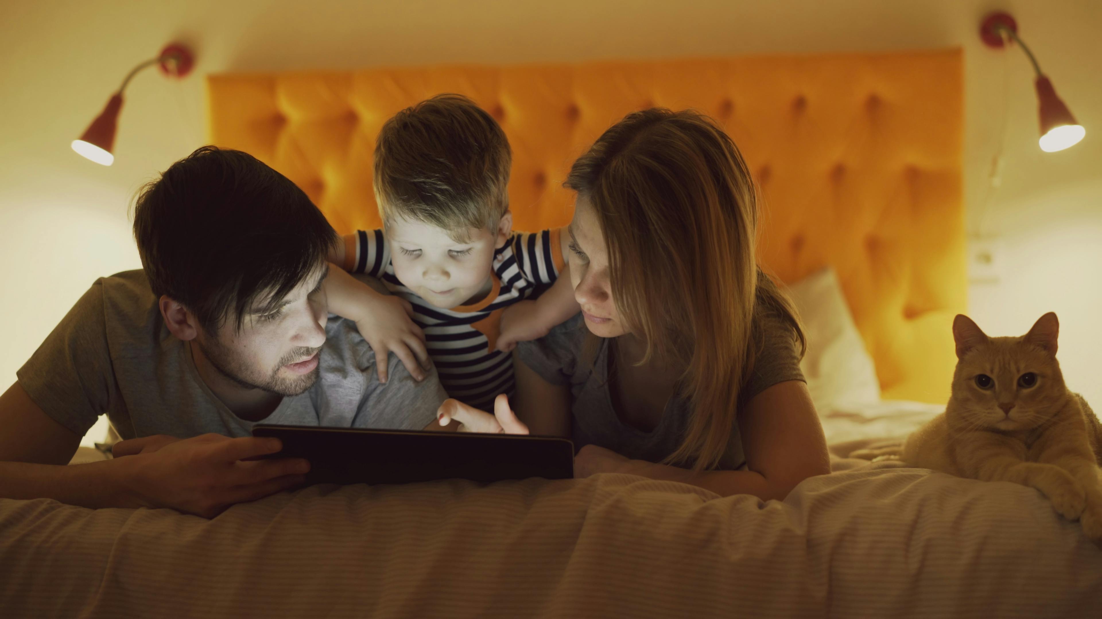 Parents and child using a tablet in bed with a cat nearby, cozy evening.