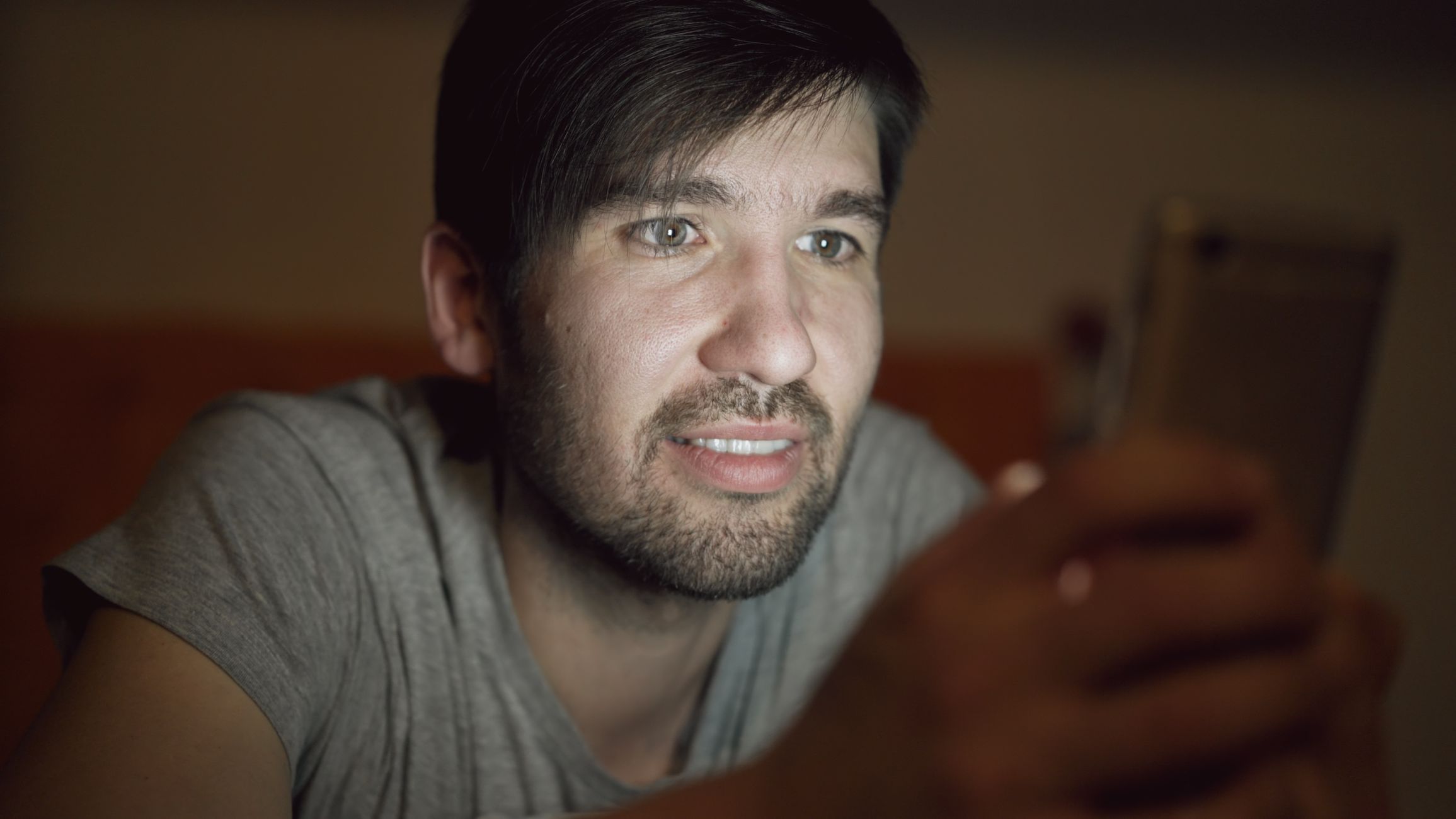 A young adult male engages with a smartphone, illuminated by its screen in a dim setting.