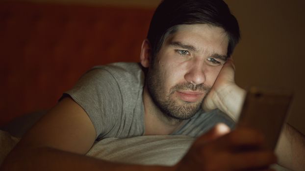 An adult man in a dim room looking at a smartphone with a thoughtful expression.
