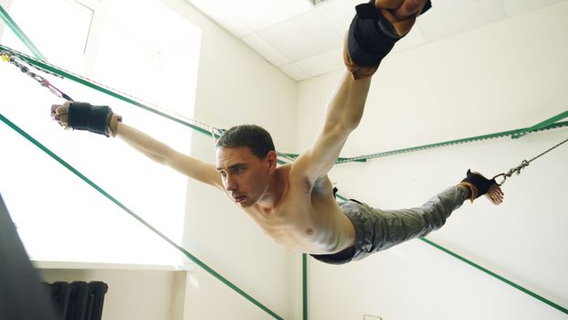 An athletic man uses suspension cables for an aerial fitness workout inside a bright room.