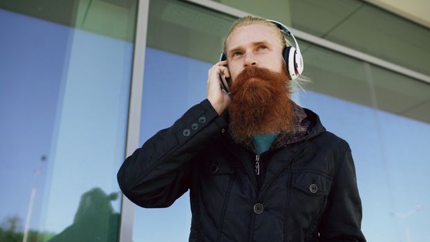 A man with a long beard wearing headphones outside a building on a sunny day.
