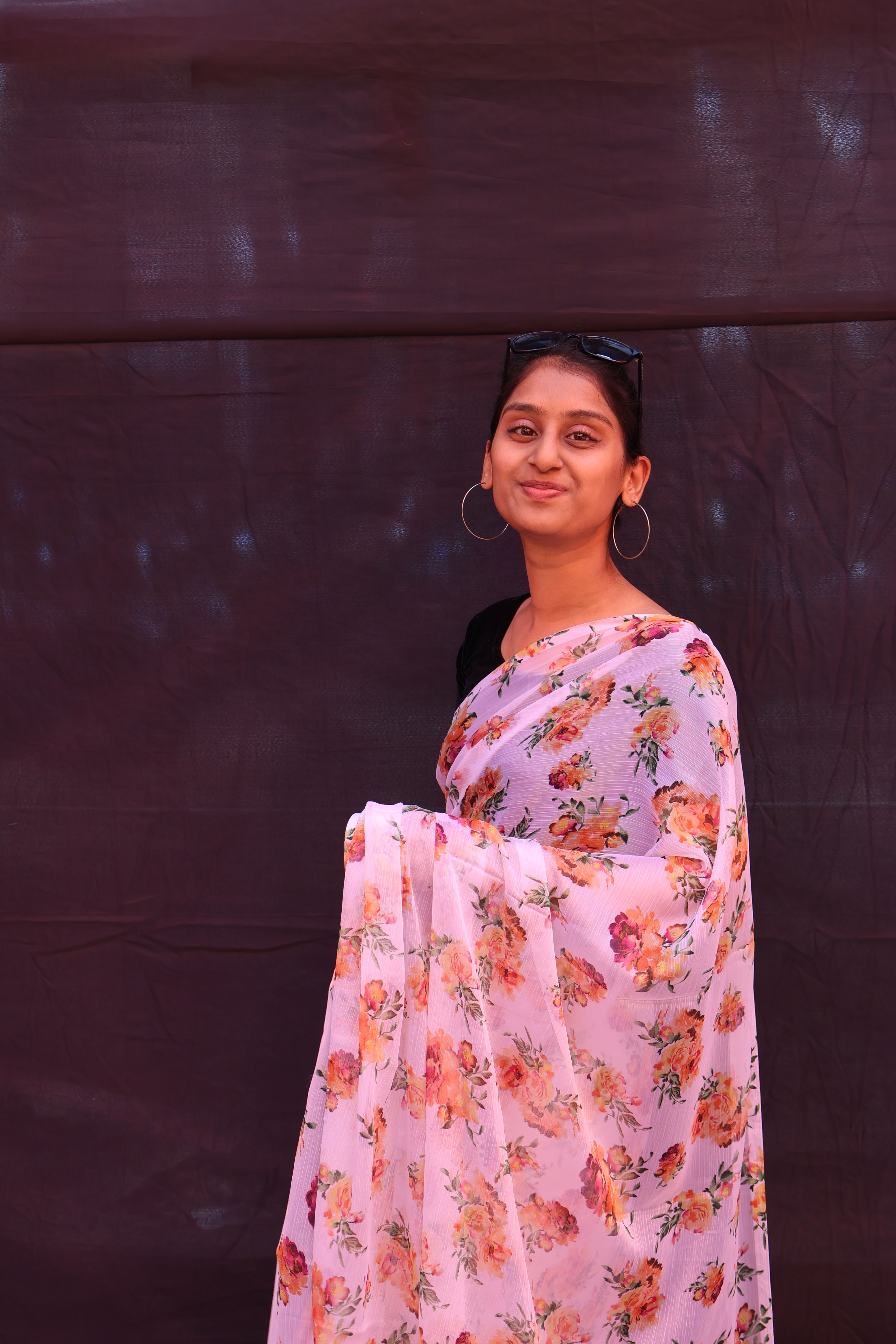 Smiling woman wearing a floral saree and hoop earrings, posing confidently against a dark backdrop.