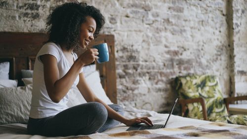 Free A woman enjoying coffee while working on a laptop in a cozy bedroom setting. Stock Photo