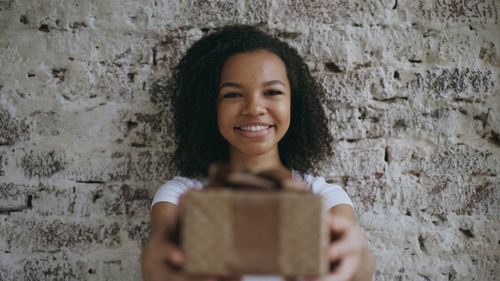 Free Smiling woman holding a gift box towards the camera against a rustic wall. Stock Photo