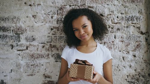Free Young woman in white shirt holding a gift box, smiling against a rustic brick wall. Stock Photo