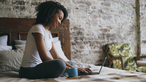 Free Young woman working on a laptop in a cozy, rustic bedroom setting. Stock Photo