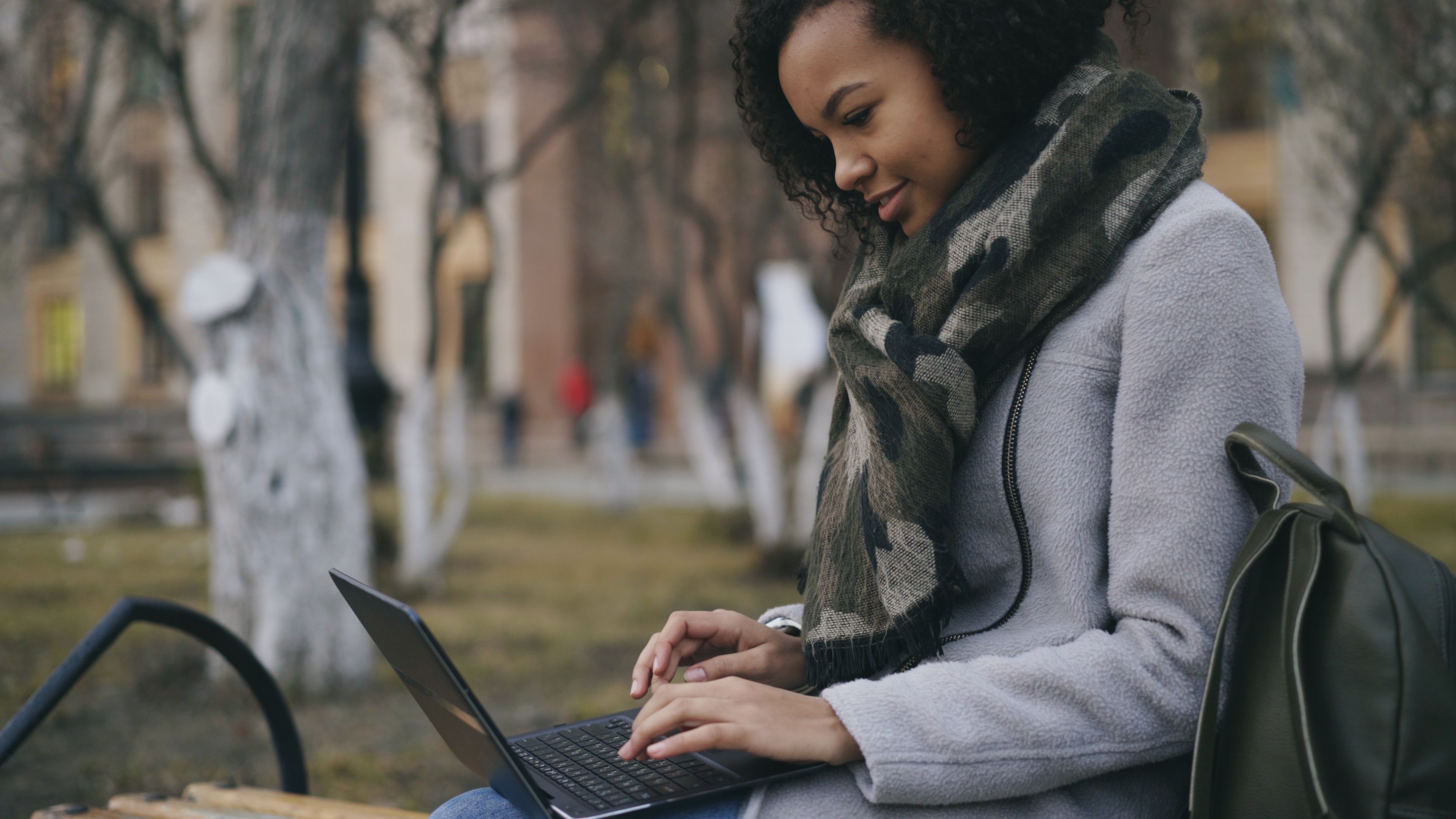 A student sitting on a park bench with a laptop, surrounded by trees and a city skyline in the background, looking focused but relaxed