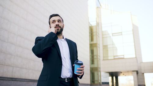 Free Businessman on phone holding a coffee cup outside a modern office building. Stock Photo