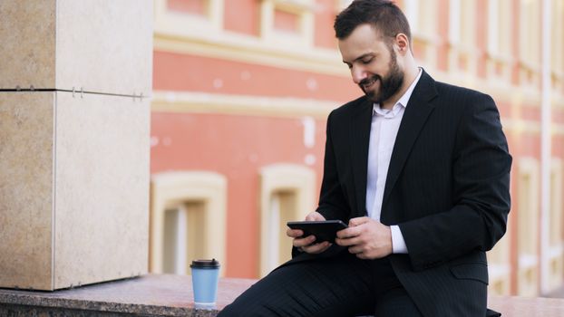 Smiling businessman sitting outdoors, using tablet with a coffee cup nearby.