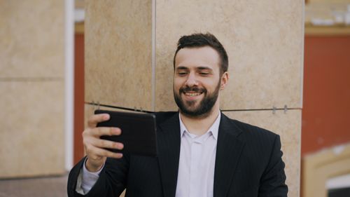 Free Smiling businessman in a suit takes a selfie with a tablet outdoors against a tiled wall. Stock Photo