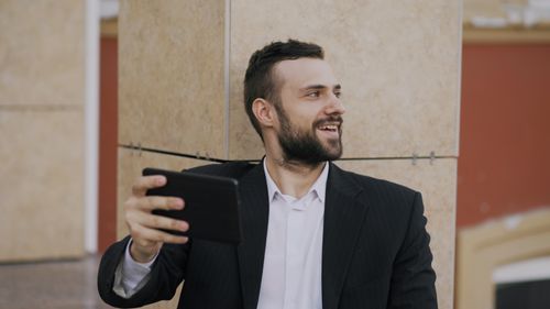 Free Smiling man in suit takes a selfie with a smartphone, sitting outdoors against a modern building. Stock Photo
