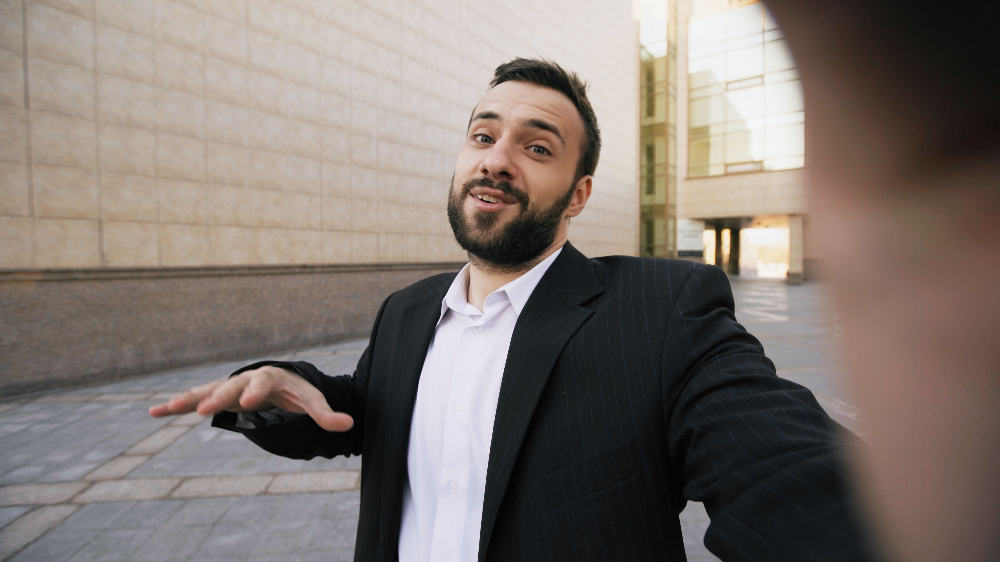 Smiling businessman in a suit taking a selfie outdoors against modern architecture.