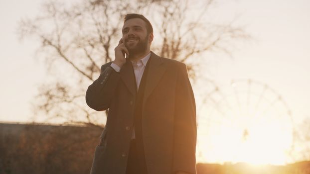 A man in a coat talking on a phone with a Ferris wheel in the sunset background.
