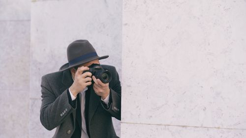 Free A person with a camera and gray hat capturing photos against a marble wall. Stock Photo