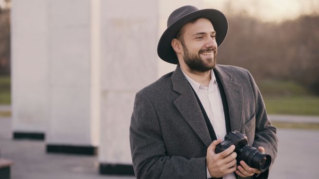 A bearded man in a coat and hat holding a camera, smiling outdoors.