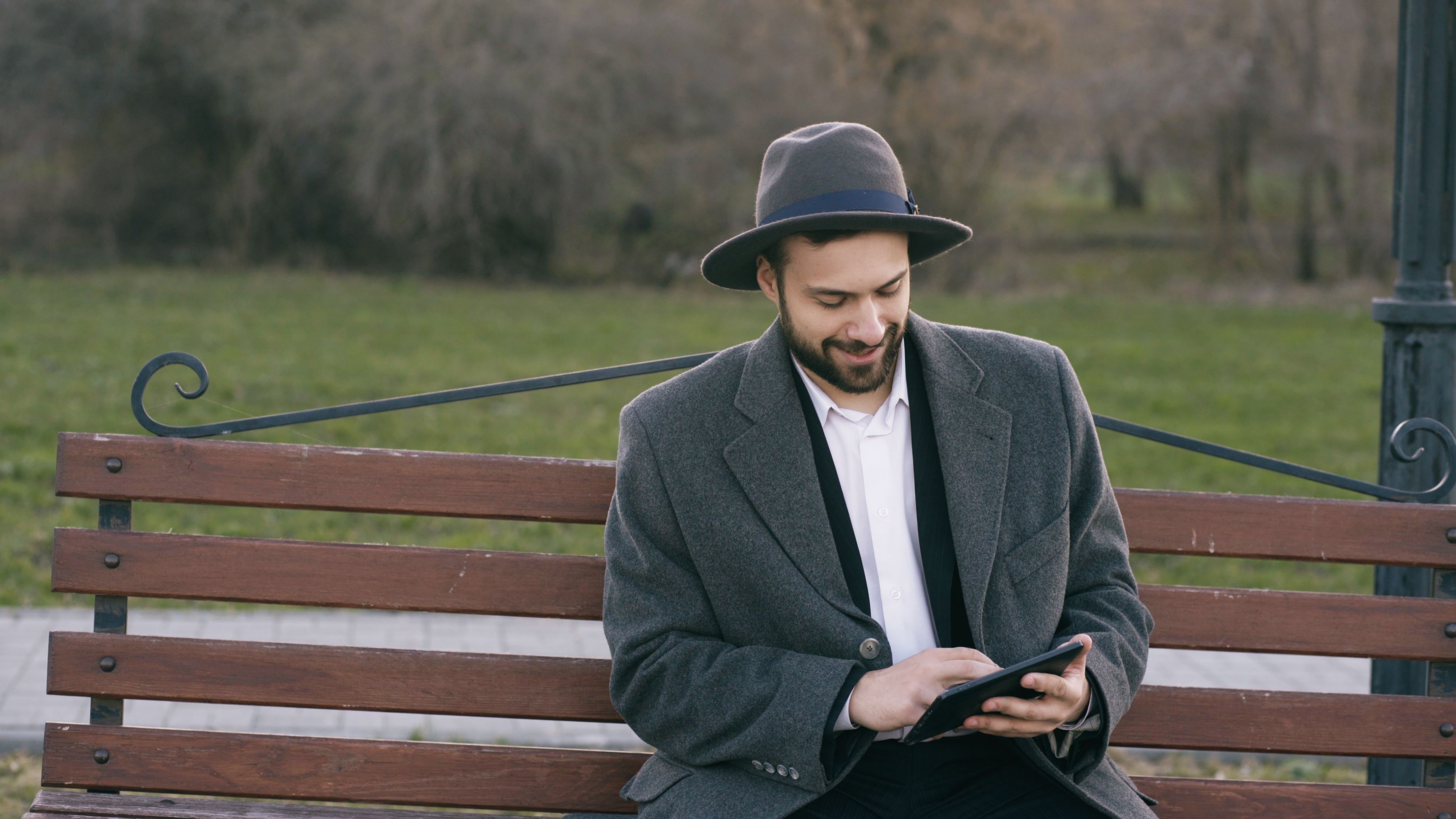 A man wearing a hat sits on a park bench using a tablet, enjoying a peaceful outdoor setting.