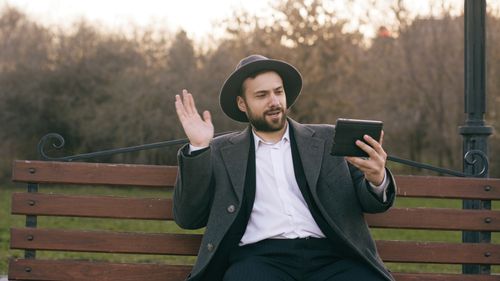 Free A bearded man in a hat video calling on a park bench during fall. Stock Photo