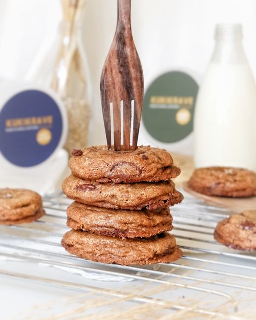 A stack of thick oatmeal cookies studded with chocolate chips and flaxseed on a wire rack, with a small dish of brewer's yeast powder beside them.