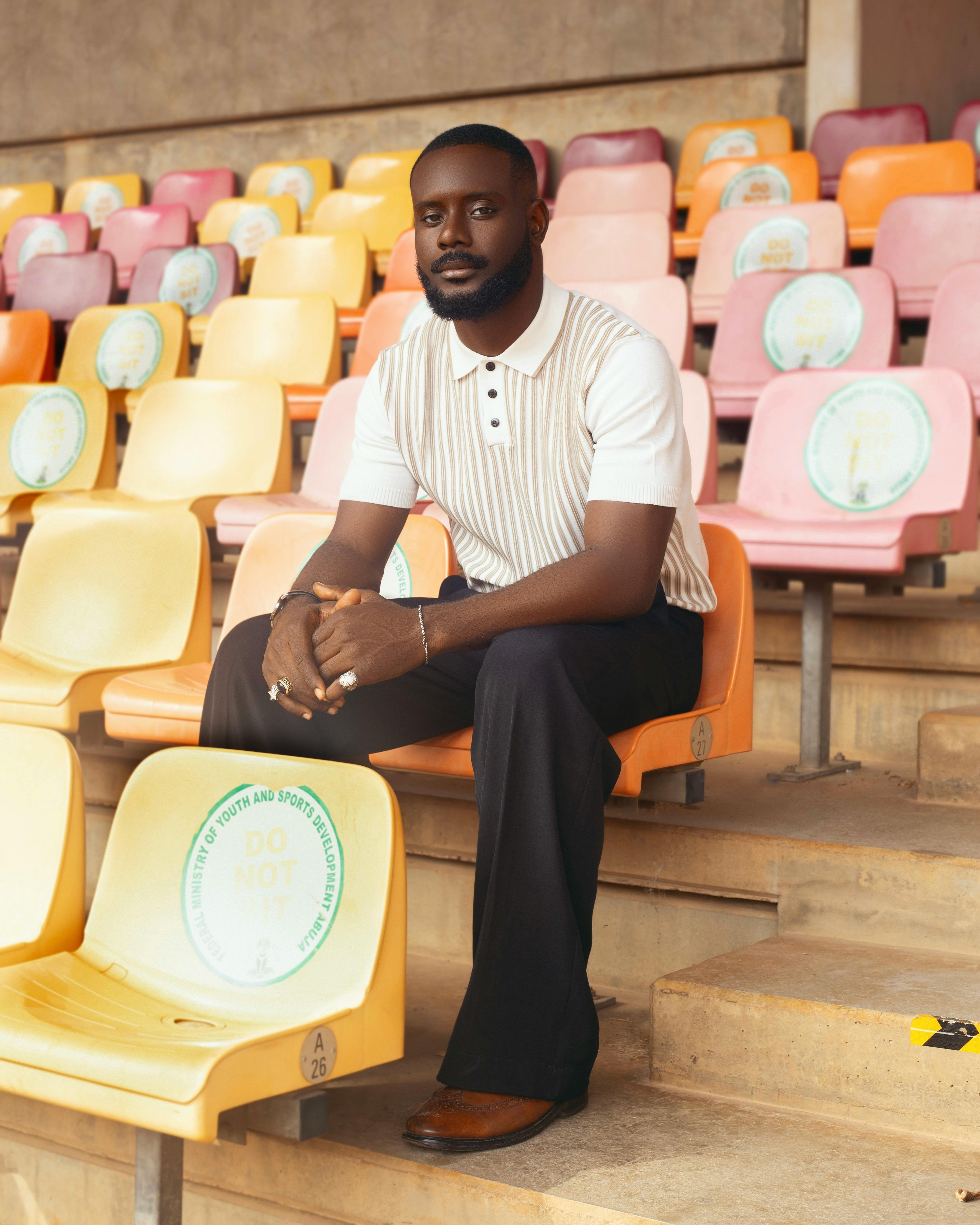 Stylish man sitting in colorful stadium seats in Abuja, Nigeria, showcasing fashion and design.
