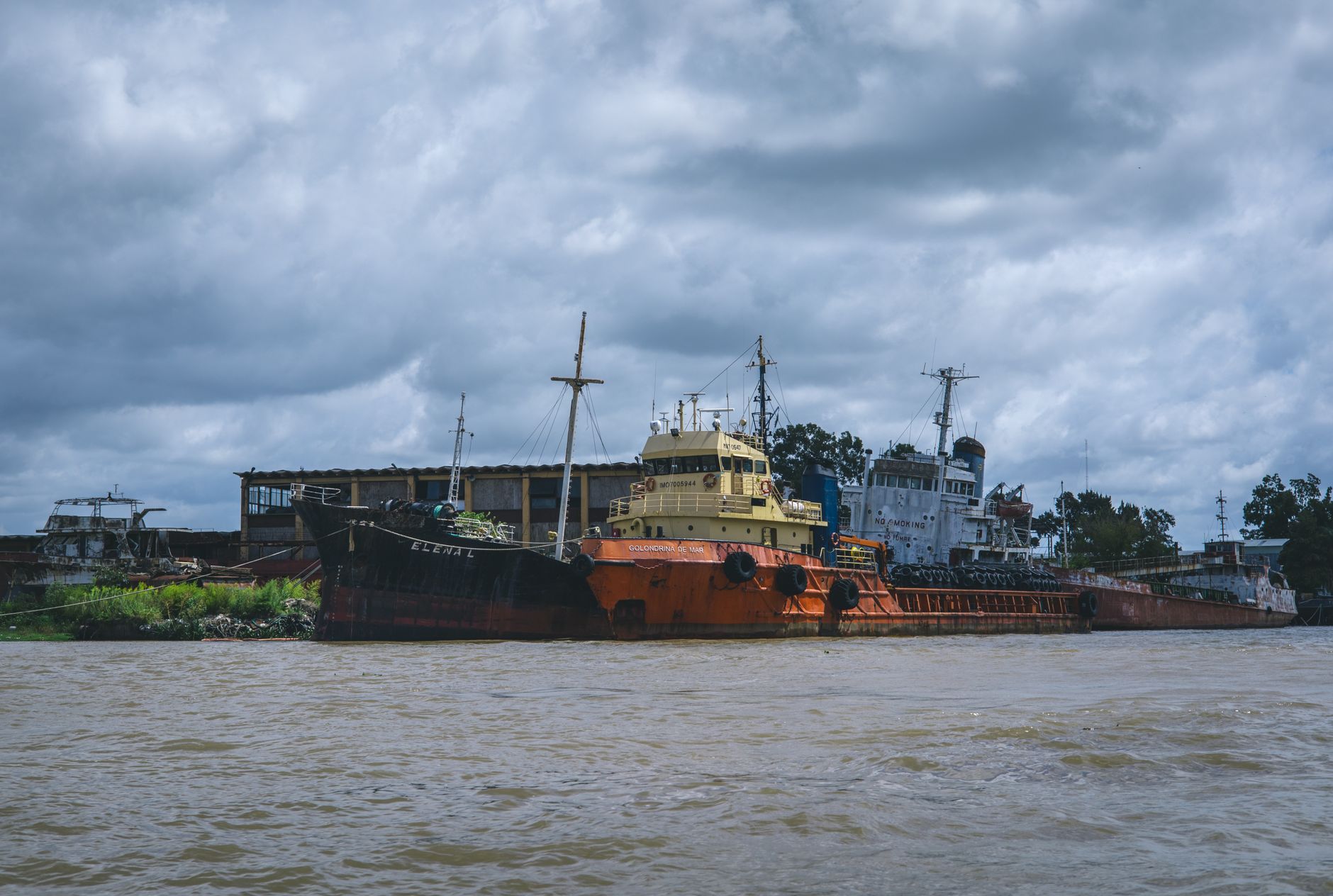 Aging cargo ships docked by the riverside under a moody, cloudy sky, showcasing maritime decay.