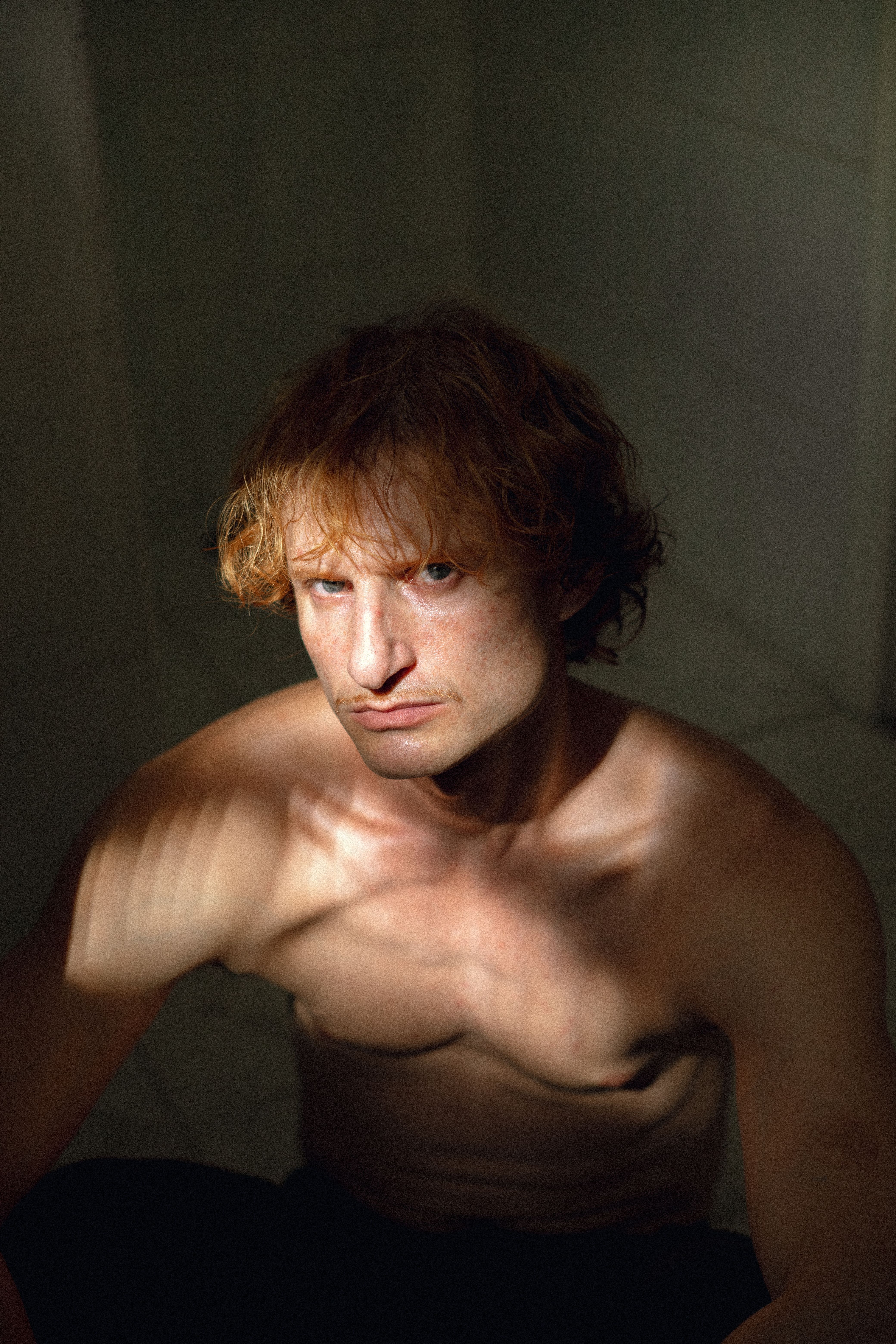A shirtless man sits in dramatic, low light creating a moody atmosphere.