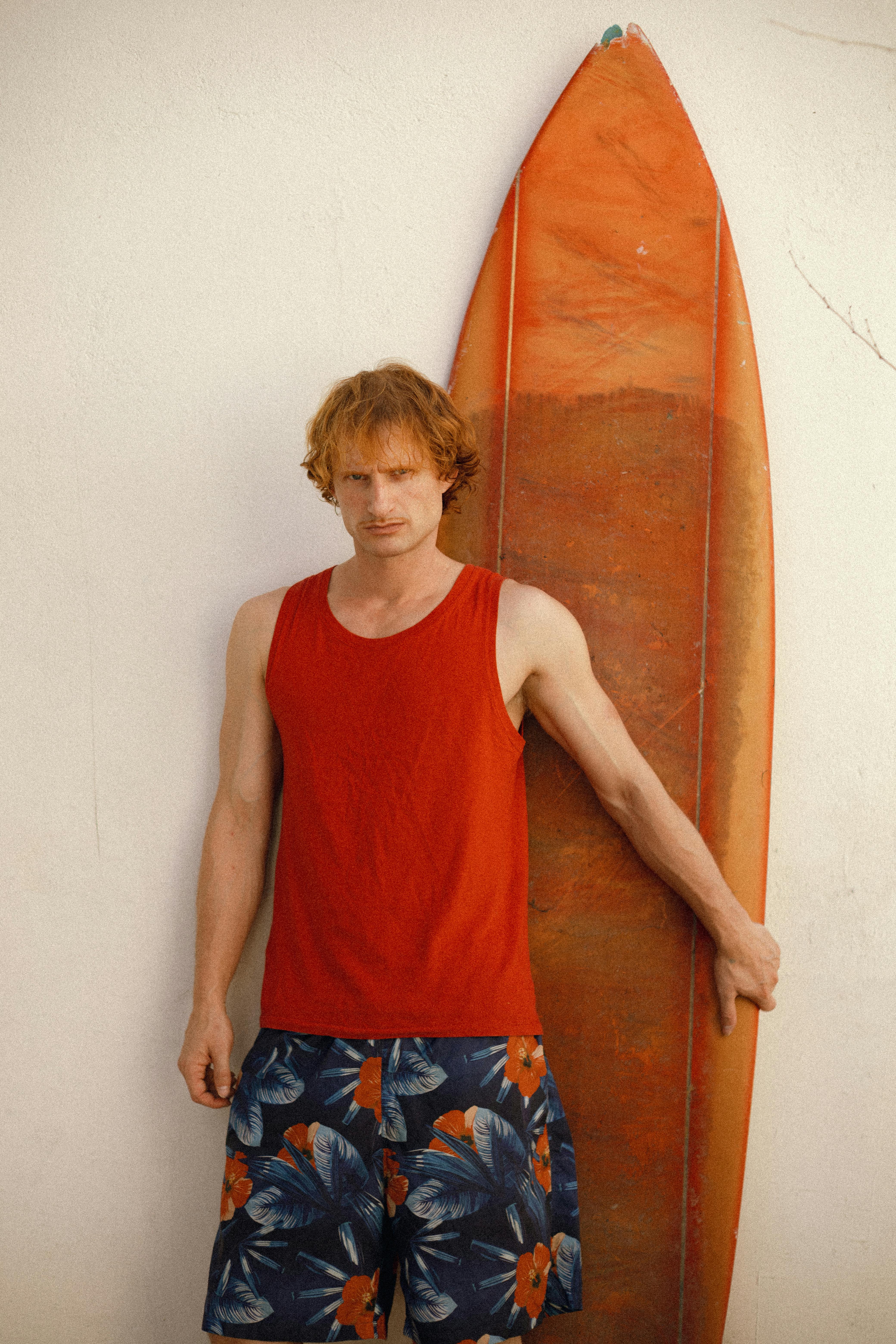 Adult male in red tank top and floral shorts with a vintage surfboard against a wall.
